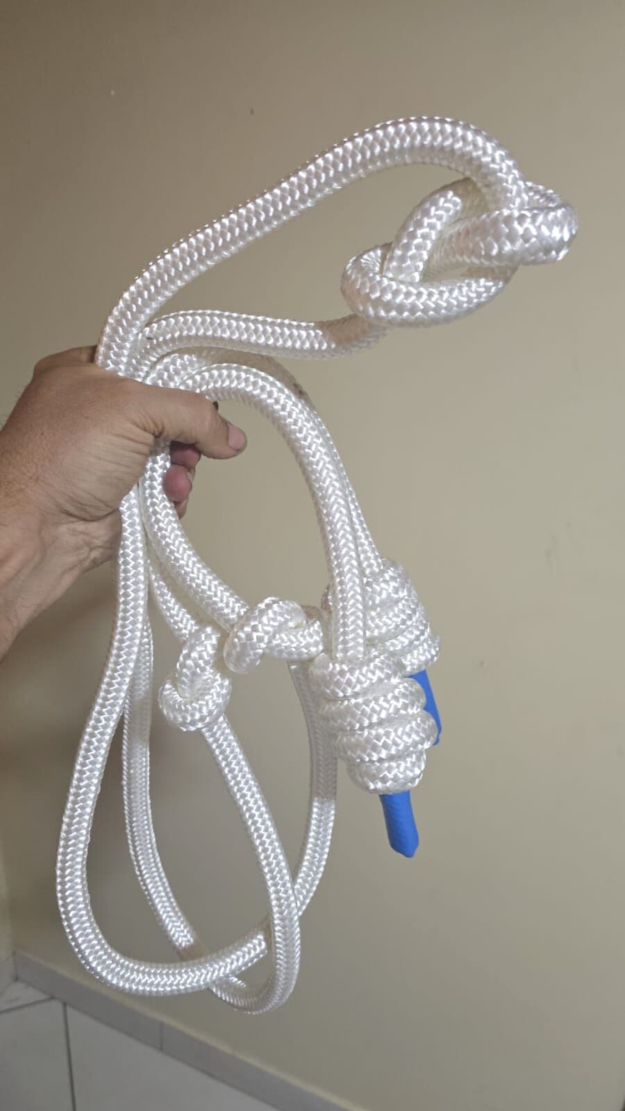 A corda branca de 16 mm segurada na mão: o nó de espirais da empunhadura, a ponteira azul na ponta e o brilho acetinado da trança.