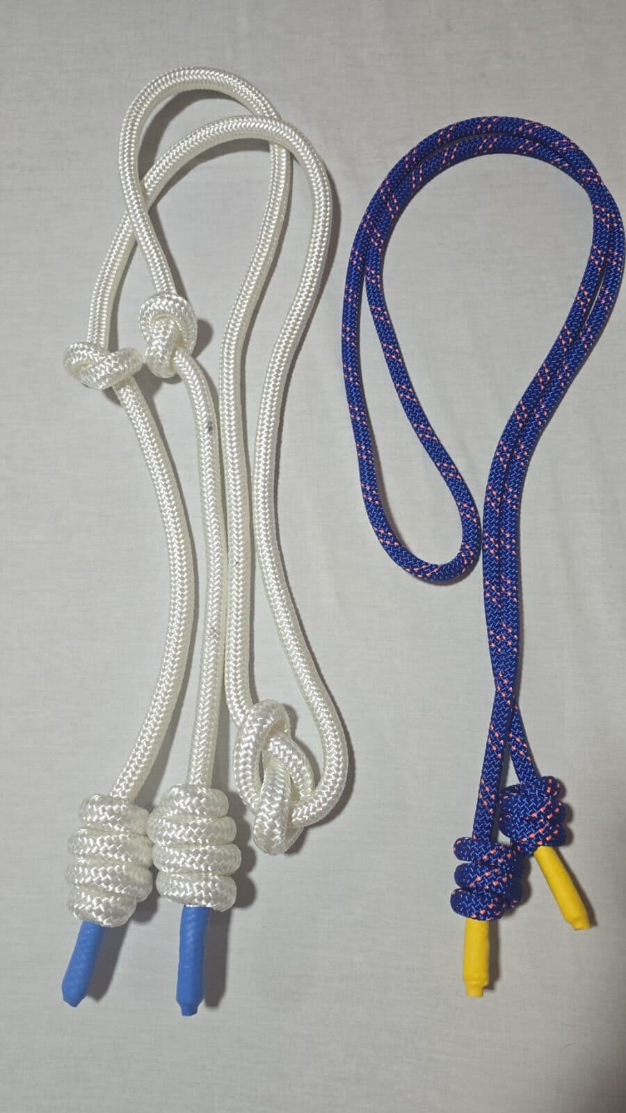 As duas cordas do kit deitadas lado a lado num tecido claro: a branca, mais grossa, com empunhaduras de nó e ponteiras azuis; a azul com fios coral, mais fina, com ponteiras amarelas.